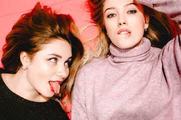 Young woman sticking out tongue looking at her girlfriend while she biting her lip. Happy lesbian couple lying on pink sofa