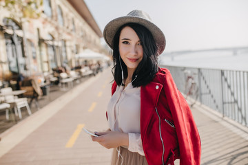 Lovely dark-haired girl in hat listening music near outdoor cafe. Photo of dreamy brunette caucasian lady in red jacket posing in earphones at embankment.