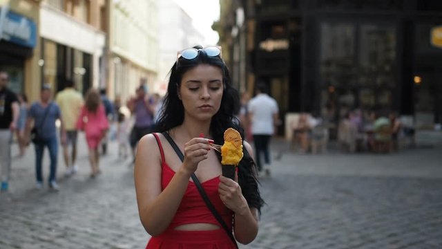 Young beauty woman in red dress is smiling and eating ice cream during walking in the street of historical part of Bratislava city in sunny summer afternoon. Half speed.