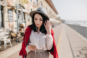 Graceful white girl looking at phone while spending time outdoor. Photo of wonderful brunette lady in red jacket walking around town in weekend.