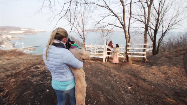 Photographer is taking photo of brunette pregnant woman stroking brown horse at photo shoot. Early spring. 