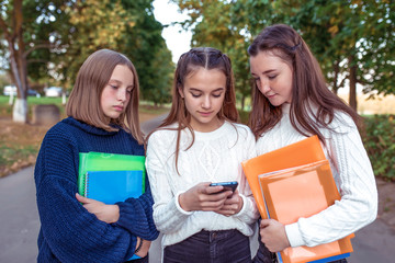 Three teenage girls schoolgirls 12-14 years old, summer city, hand smartphone, online Internet, social networks, watching photos Internet. Break school college. In hands notebooks folders textbooks.