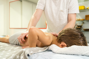 Obraz na płótnie Canvas Small caucasian boy laying on the bed naked having back massage by professional female physiotherapist masseuse in a rehabilitation center salon or home office of physical therapy side view