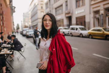 Obraz premium Pleased caucasian girl in glasses standing on the street with phone in hand. Smiling brunette young lady in red jacket posing with pleasure in city.