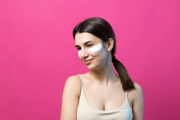 Close up portrait of pretty attractive girl with naked shoulders using patches under eyes. Standing over pink background.