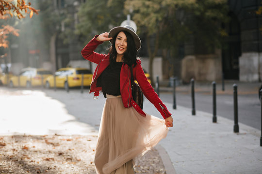 Wonderful brunette girl with charming smile playing with long skirt on city background. Outdoor photo of beautiful caucasian lady in hat relaxing in autumn park.