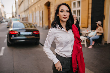Confident caucasian girl with black hair posing on the street with car on background. Outdoor photo of confident female office worker relaxing after work.