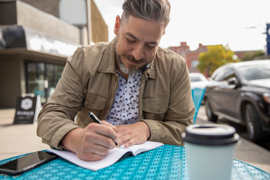 Man Working, Writing In Notebook At Sidewalk Cafe