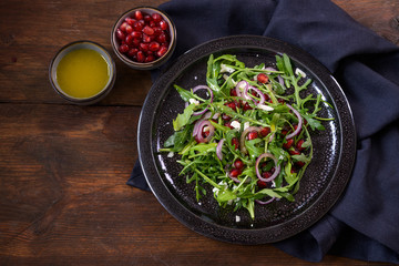 Arugula or rocket salad with pomegranate, red onions and parmesan served on a dark plate, small bowls with seeds and dressing, rustic wooden background, copy space, high angle view from above