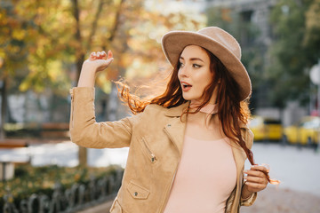 Elegant ginger girl in beige jacket looking away with surprised face expression. Outdoor photo of red-haired woman in hat expressing amazement on nature background.