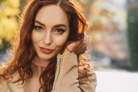 Close-up portrait of sensual young woman playing with dark ginger hair. Dreamy red-haired girl posing with smile on blur background.