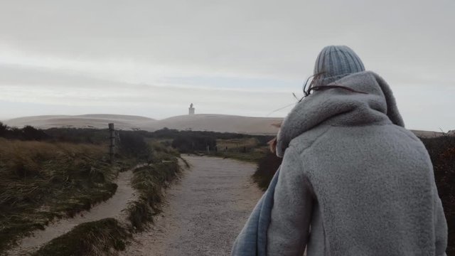 Camera follows thoughtful tourist woman in winter clothes walking towards far away overcast beach lighthouse slow motion