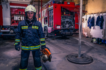 A fireman with uniform and helmet holding a chainsaw with fire truck in the background