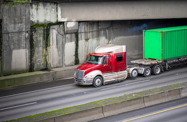 Big rig low cab semi truck transporting green container on flat bed semi trailer running on the road overpass intersection and bridge