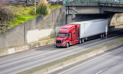 Big rig red bonnet long haul semi truck transporting cargo in dry van semi trailer running under the bridge over the wide divided highway