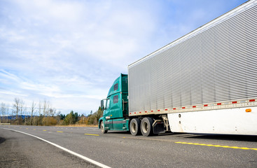 Big rig semi truck with corrugated refrigerated semi trailer with aerodynamic skirt moving on the road with trees on the side