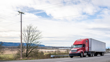 Red big rig semi truck transporting cargo in dry van semi trailer running on the winding road with trees and meadow