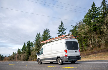 Small compact commercial cargo mini van with ladders on the roof running on the road with trees on the hill. © vit