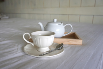 White color ceramic coffee cup with white color teapot on timber trey on white bed sheet