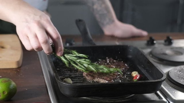 a male cook roasts a piece of marble steak in a grill pan. the chef spreads a sprig of rosemary on a fresh piece of beef in the restaurant's kitchen. Grilled steak in a restaurant. Cooking meat with s