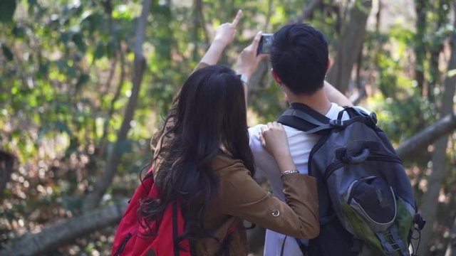 HD Blurred Slow Motion rear view of happy young Asian couple man and beautiful woman holding hands together and hiking with backpack in forest mountain with using smartphone taking a photo of nature.