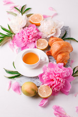 Breakfast for Valentines day with cup of tea, peony flowers and croissant on gray murble table and white background from above, flat lay, wedding, birthday, romance concept