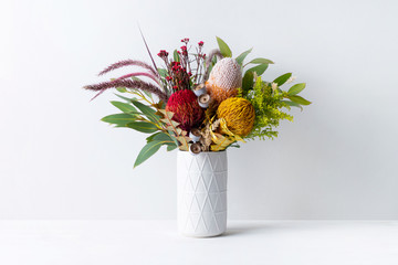 Beautiful floral arrangement of mostly Australian native banksias, eucalyptus leaves and gum nuts, fountain grass and dried kangaroo grass, in a white vase on a white table with a white background.