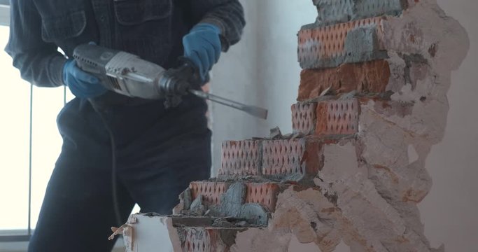 Male builder work with jackhammer, demolishes wall in apartment, pick out bricks from masonry, breaks piece, throws it to side, cleans dust and debris, stones fly in different directions, construction