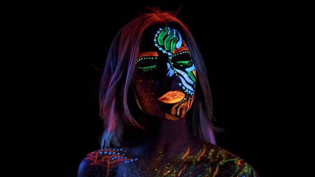 Closeup of Expressive Colored Painted Female Face. Neon Pigment on Skin. Shiny Contrast Paints Glowing in Fluorescent Light. Girl Rise Up Head and Looks in Camera. Sparkle Eyes, Orange Lips.