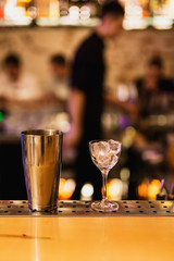 A nick and nora glass filled with ice and a shaker on a bar counter, people in bokeh, bar atmosphere