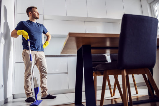 Full length of handsome tired man standing in kitchen and leaning on mop. There is so much to clean. - Powered by Adobe