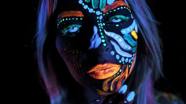Close-up of Impressive Fashion Girl with Ultraviolet Bright Eye Lenses Looking in to the Camera. Face with Ultra Violet or Lumenescent Makeup. Colored Brows, Lips, Pink Hair. Black Background.