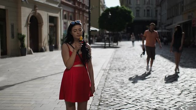 Young beauty woman in red dress is smiling and eating ice cream during walking in the street of historical part of Bratislava city in sunny summer afternoon. Half speed.  Close up.
