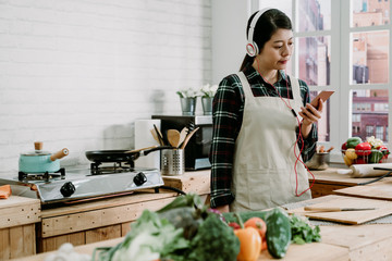 Portrait of happy young asian korean lady listening to music from headphones while cooking in kitchen. wife looking at mobile phone and smiling while enjoy songs in earphones during prepare lunch
