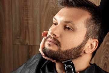 Obraz premium The master makes a beard haircut to the client with an electric machine, trimmer, close-up. The process of shaving a beard at the hairdresser. Body care, lifestyle, metrosexual.