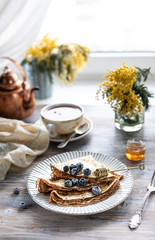 A plate with pancakes with blueberry berries on a wooden table. In the background is a cup of tea and a bouquet of spring flowers by the window.