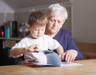 Portrait of beautiful mature woman (80 years old) with her great-grandson at home, reading educational book together