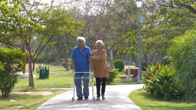 Happy old couple enjoying a Sunday morning walk in a hospital park - lifestyle couples. Senior white-haired man walking slowly with the help of a walker while his wife is assisting him - nursing an...
