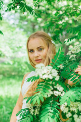 Portrait of a charming blond woman wearing beautiful white dress standing next to rowan tree with white flowers.