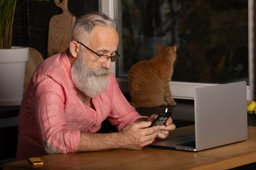Handsome bearded old man dressed in smart casual style smoking and using a smartphone at home