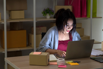 Startup small business entrepreneur young Asian woman small business owner sitting in fron of laptop on wooden table checking sales results at home at night