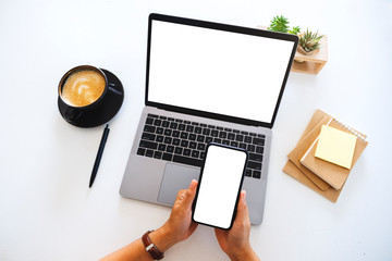 Top view mockup image of hands holding a blank white screen mobile phone and laptop computer on the...
