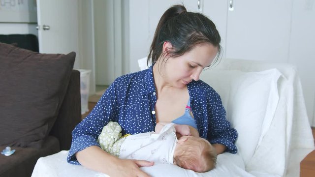 Proud mother with maternity bra breastfeeds her latching baby on comfy chair while stroking their back