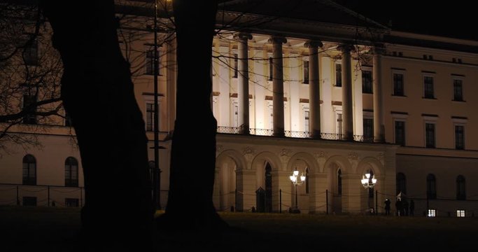 Stabilized wide 4K shot of dark black trees moving from right to left in parallax motion in front of illuminated Norwegian Royal Palace building with iconic balcony, at night in Oslo Norway.