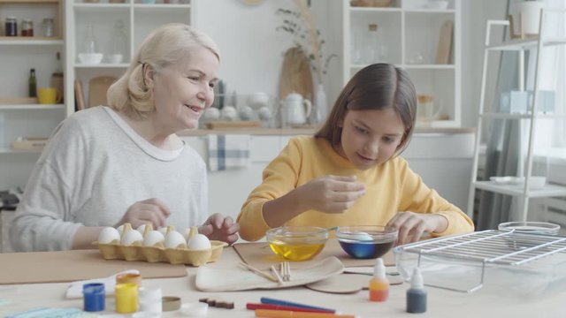 Cute girl dipping egg into bowl with blue food coloring and dying it with help of joyous grandmother while preparing for Easter at home