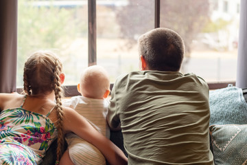 A blond girl, her baby brotherand their father looking through the window. Family concept