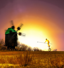 a clown on a unicycle riding with a lance in his hand towards the windmil during sunset. © Eziz