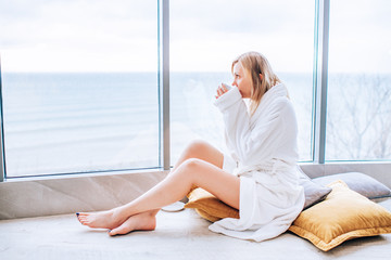 Woman in a white bathrobe enjoying sea view next to big window with cup of coffee. Panoramic window. Floor covering, Vacation in a hotel
