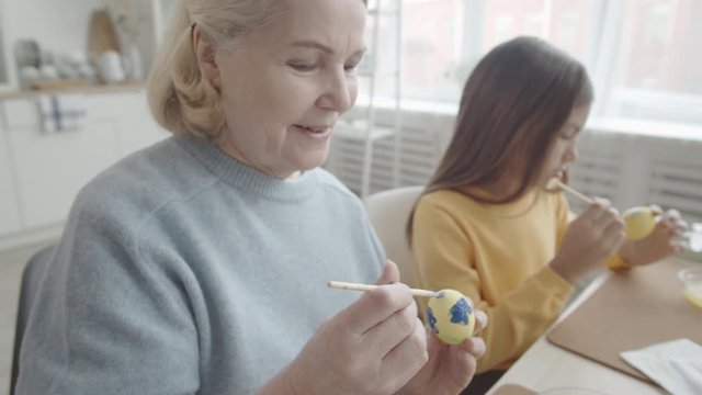 Arc shot of cheerful senior grandmother and her little granddaughter decorating dyed Easter eggs with brush and gouache while sitting together at kitchen table