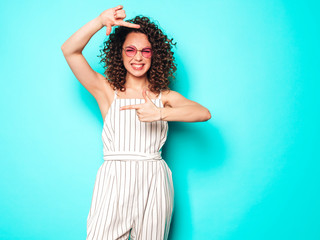 Portrait of beautiful smiling model with afro curls hairstyle dressed in summer hipster clothes.Sexy carefree girl posing near blue wall.Trendy woman makes photo frame by hands